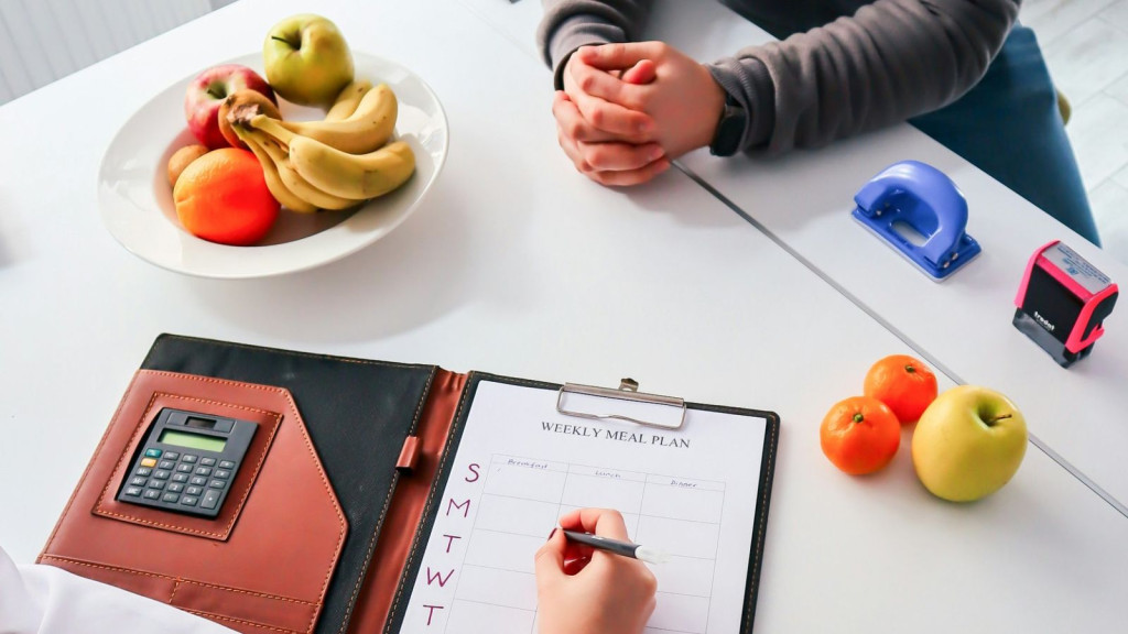 Dietista-nutricionista preparando un plan semanal de comidas durante una consulta con un paciente, con un plato de fruta sobre la mesa